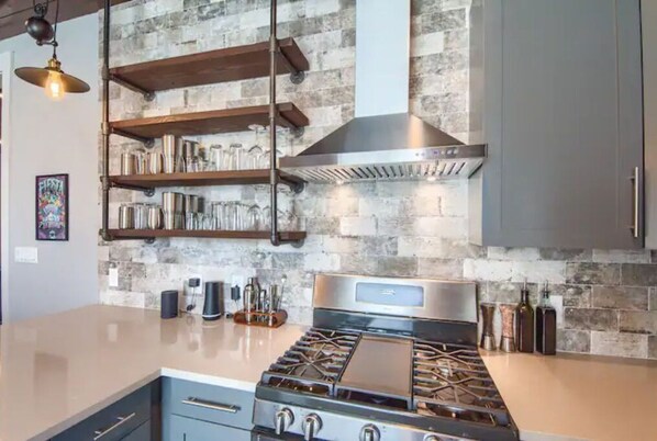 A kitchen with a stainless steel stove, wooden shelves, and a stone backsplash.