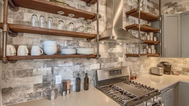 A modern kitchen with exposed brick walls, wooden shelves, and a stainless steel stove.