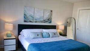 Ein ordentlich bezogenes Bett mit blauer Decke, zwei Nachttische mit Lampen, ein großes abstraktes Gemälde über dem Bett und ein Spiegel vom Fußboden bis zur Decke an der Wand.