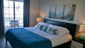 Ein ordentlich bezogenes Bett mit blauer Decke, einem Kopfteil und einem Bild darüber. Ein Nachttisch mit Lampe und Schublade ist ebenfalls zu sehen.
