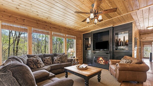 A cozy cabin interior with a fireplace, wooden furniture, and large windows overlooking a forest. - A Blessing An Upscale Mountain Getaway (Bryson City)