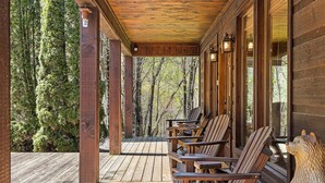 A wooden deck with chairs and a bear sculpture, surrounded by tall trees. - A Blessing An Upscale Mountain Getaway (Bryson City)