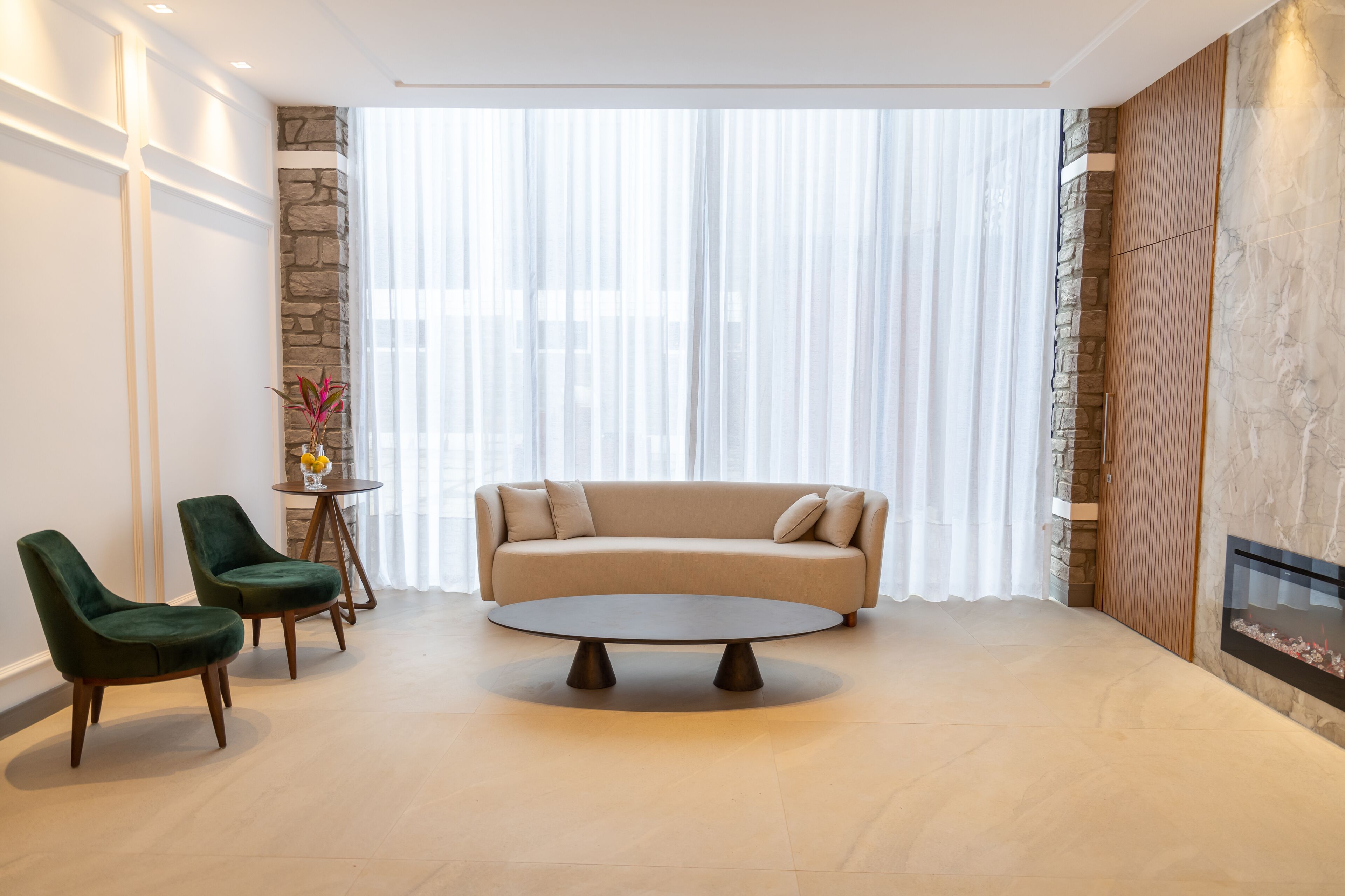 Una sala de estar moderna con un sofá color beige, dos sillones verdes, una mesa redonda y una pared de acento de piedra.