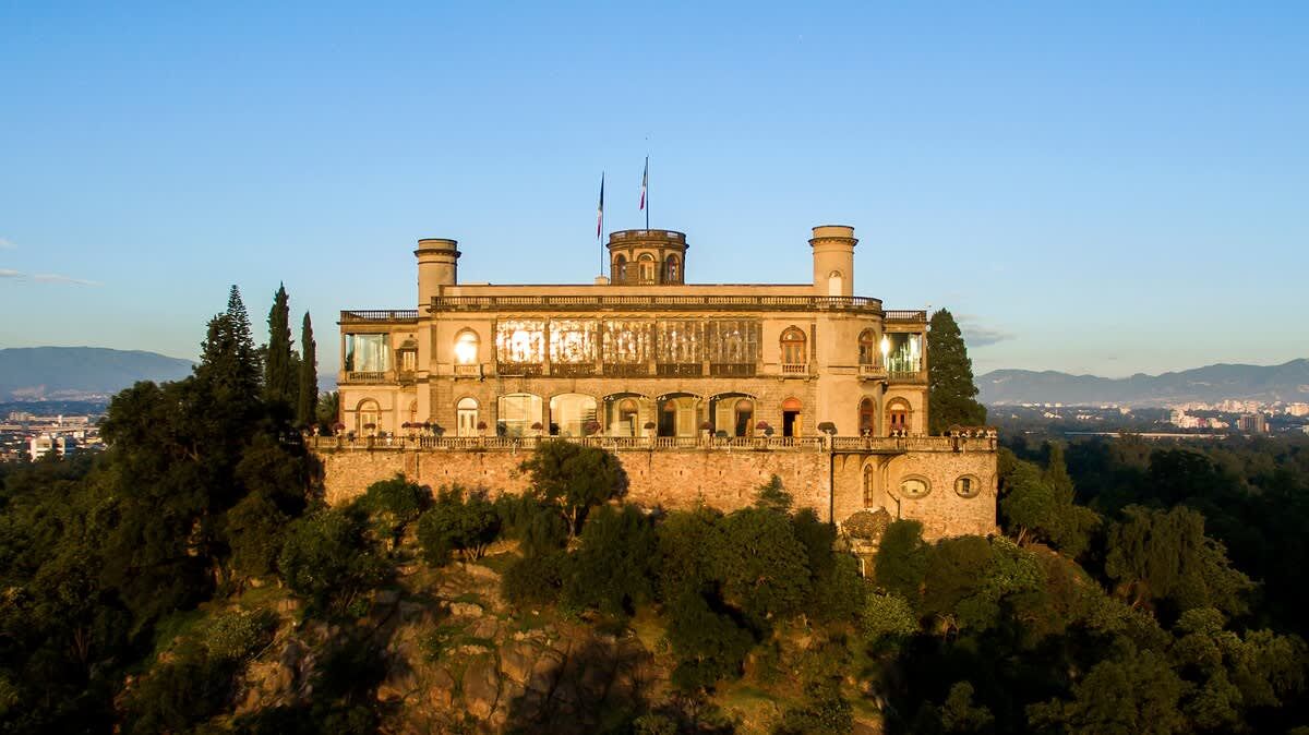 A historic building with multiple arches and a tower, surrounded by trees and overlooking a cityscape.