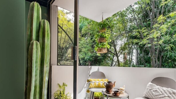 Een balkon met een cactus, planten in potten, een tafel met een theepot en een stoel.