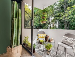 A balcony with a cactus, potted plants, a table with a teapot, and a chair.
