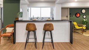 Een moderne keuken met een centraal kookeiland, twee barstoelen en een eethoek met een gele stoel.