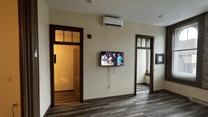 Un pasillo con una televisión de pantalla plana montada en la pared, piso de madera y una ventana con persianas.