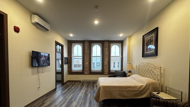 Un dormitorio con cama, una silla, un televisor y ventanas amplias.