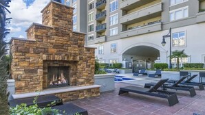 Una chimenea de piedra con fuego, zona de estar exterior, piscina y un edificio de varios pisos con balcones.