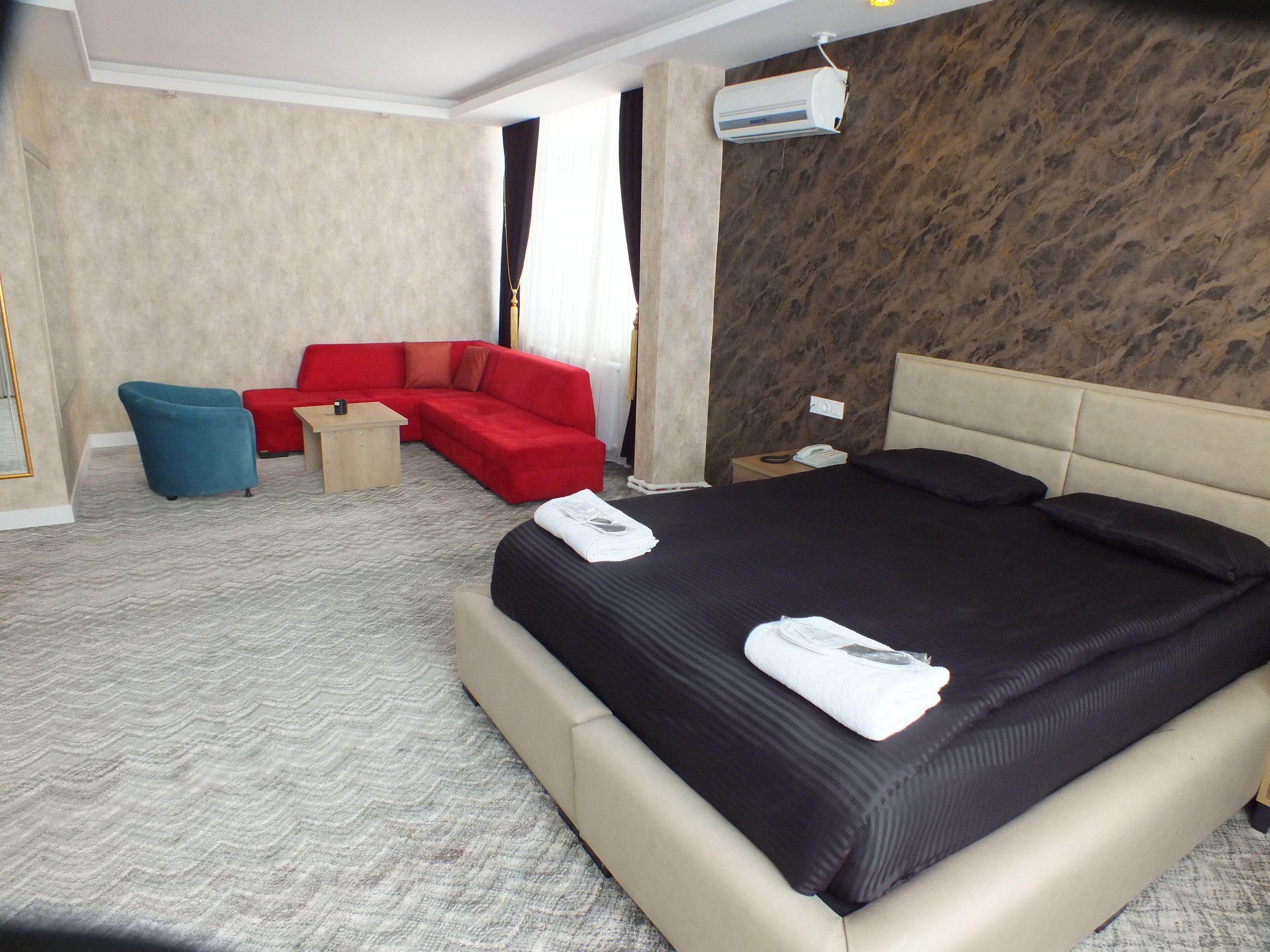 A hotel room with a large bed, a red sofa, a blue armchair, a small table, and an air conditioning unit on the wall.