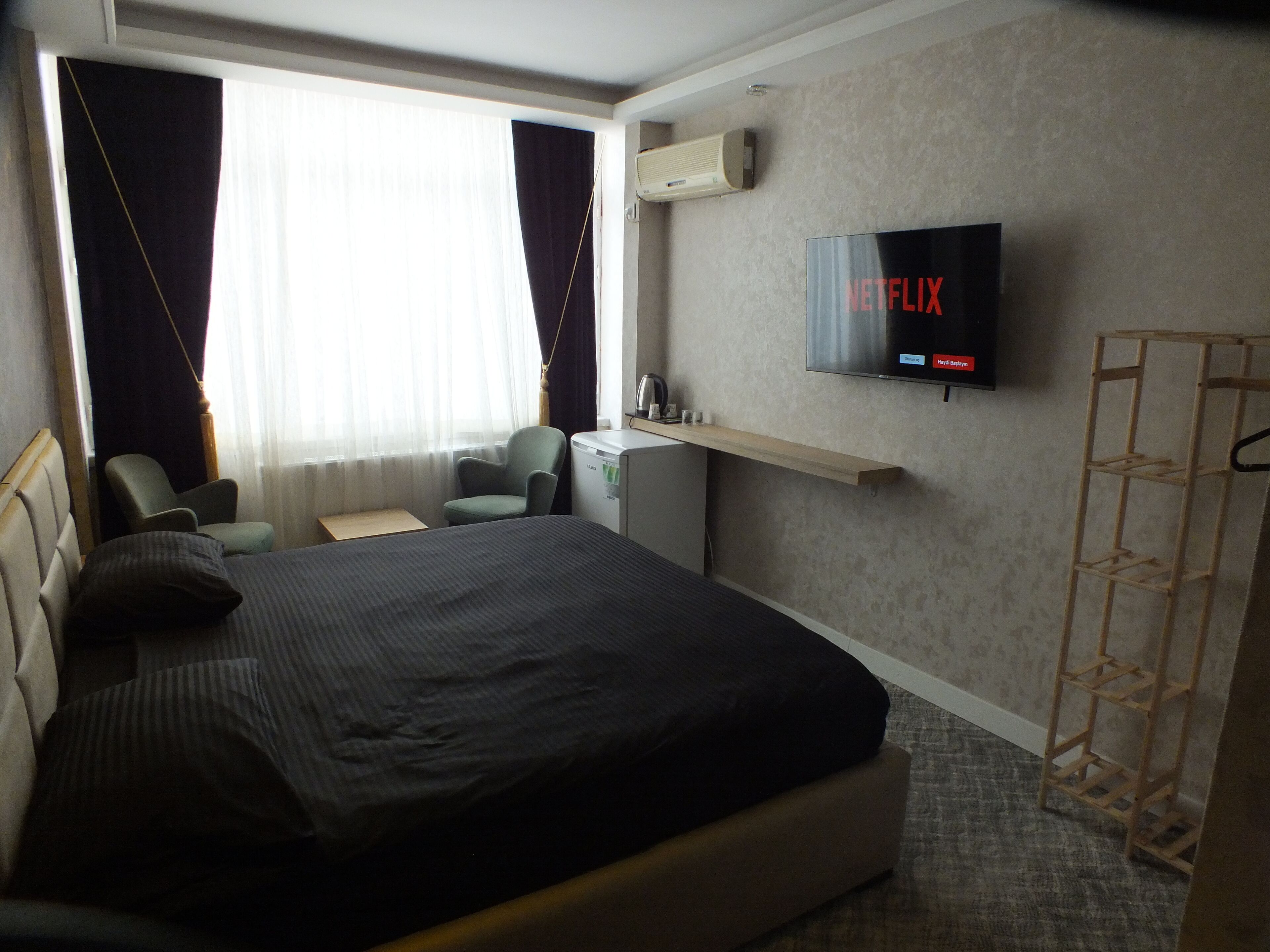 Habitación de hotel con cama, escritorio, silla, televisor montado en la pared y ventana con cortinas.