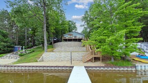 A house with a dock leading to a pond, surrounded by trees and a white fence.