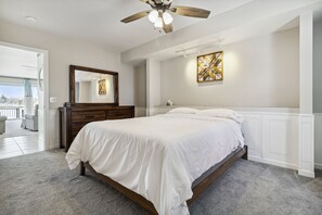 A bedroom with a large bed, a ceiling fan, a dresser, a mirror, and a wall art piece.