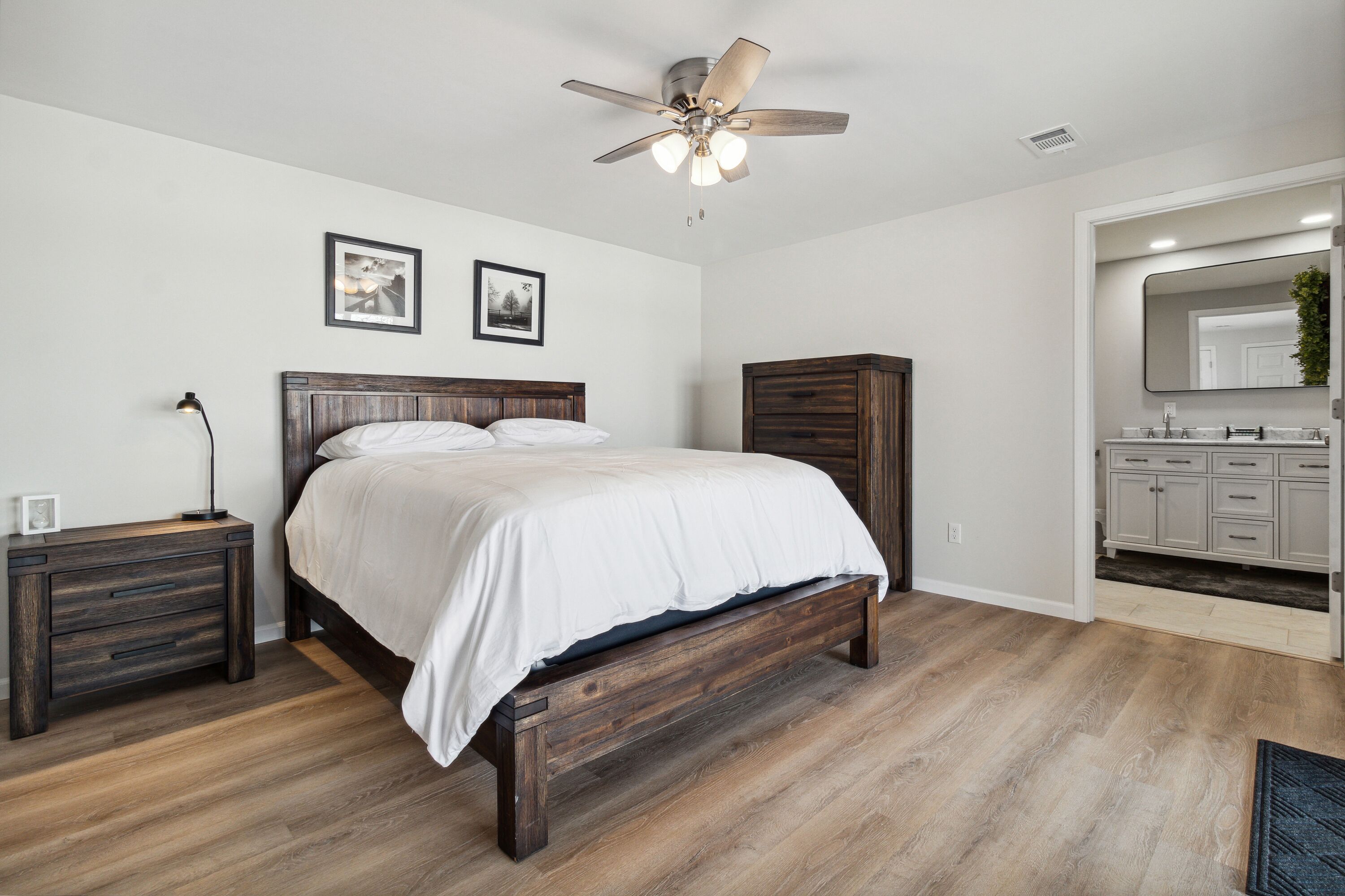 A bedroom with a wooden bed, a nightstand, a ceiling fan, and a bathroom with a sink and cabinets.