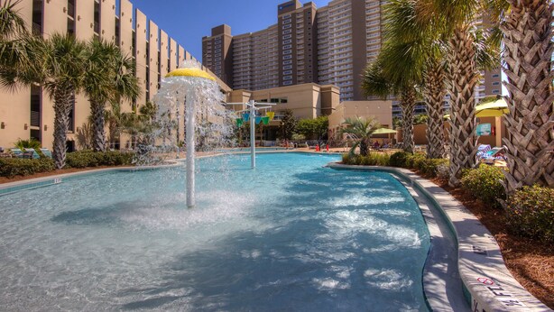 A swimming pool with a water fountain, surrounded by palm trees and a hotel building in the background.