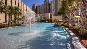 Una piscina con una fuente de agua, rodeada de palmeras y un edificio de hotel al fondo.
