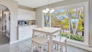 A kitchen with white cabinets, a microwave, and a dining table with chairs. There is a large window with a view of trees and a garden.