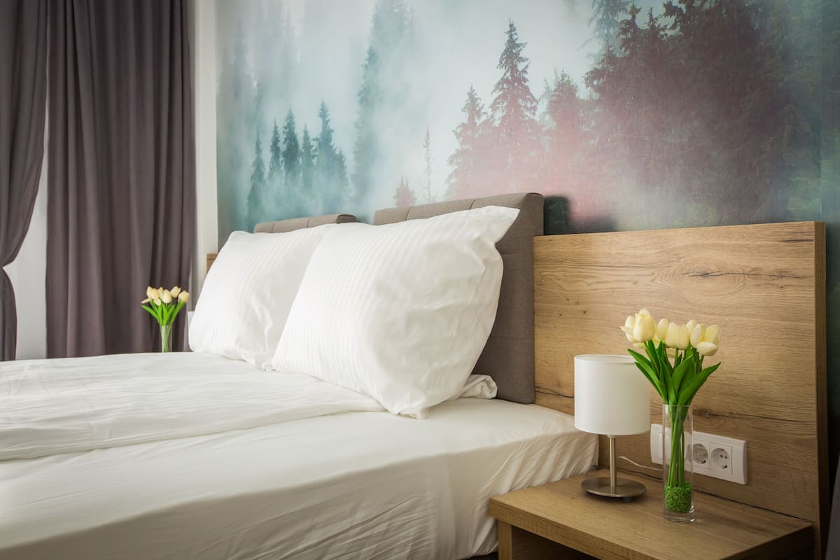 Hotellihuone, jossa on sänky, tyynyjä, yöpöytä lampun kanssa ja maljakko kukkineen.