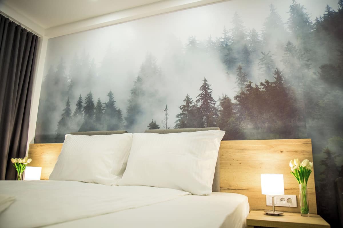 Hotellihuone, jossa on sänky, kaksi yöpöytää, yölamppu, seinämaalaus, jossa on kuvattuna metsä, ja maljakko kukkineen.