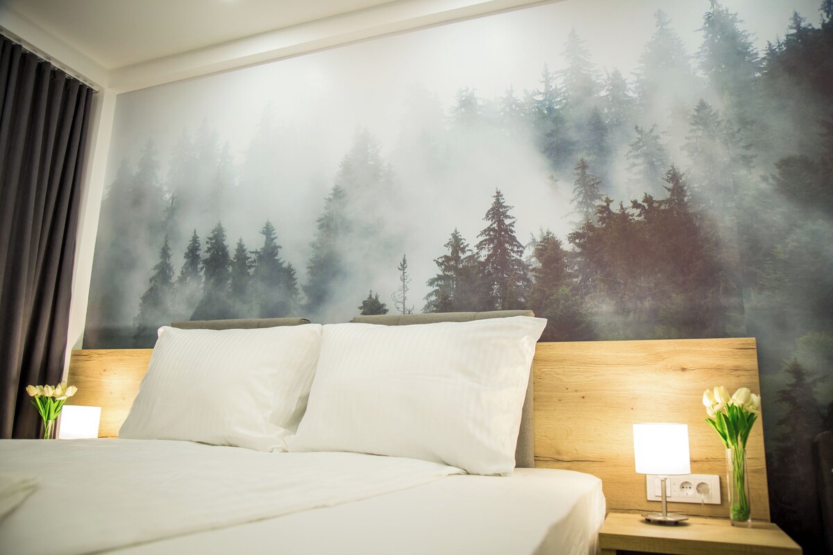 A hotel room with a bed, two bedside tables, a nightstand, a wall mural of a forest, and a vase with flowers.