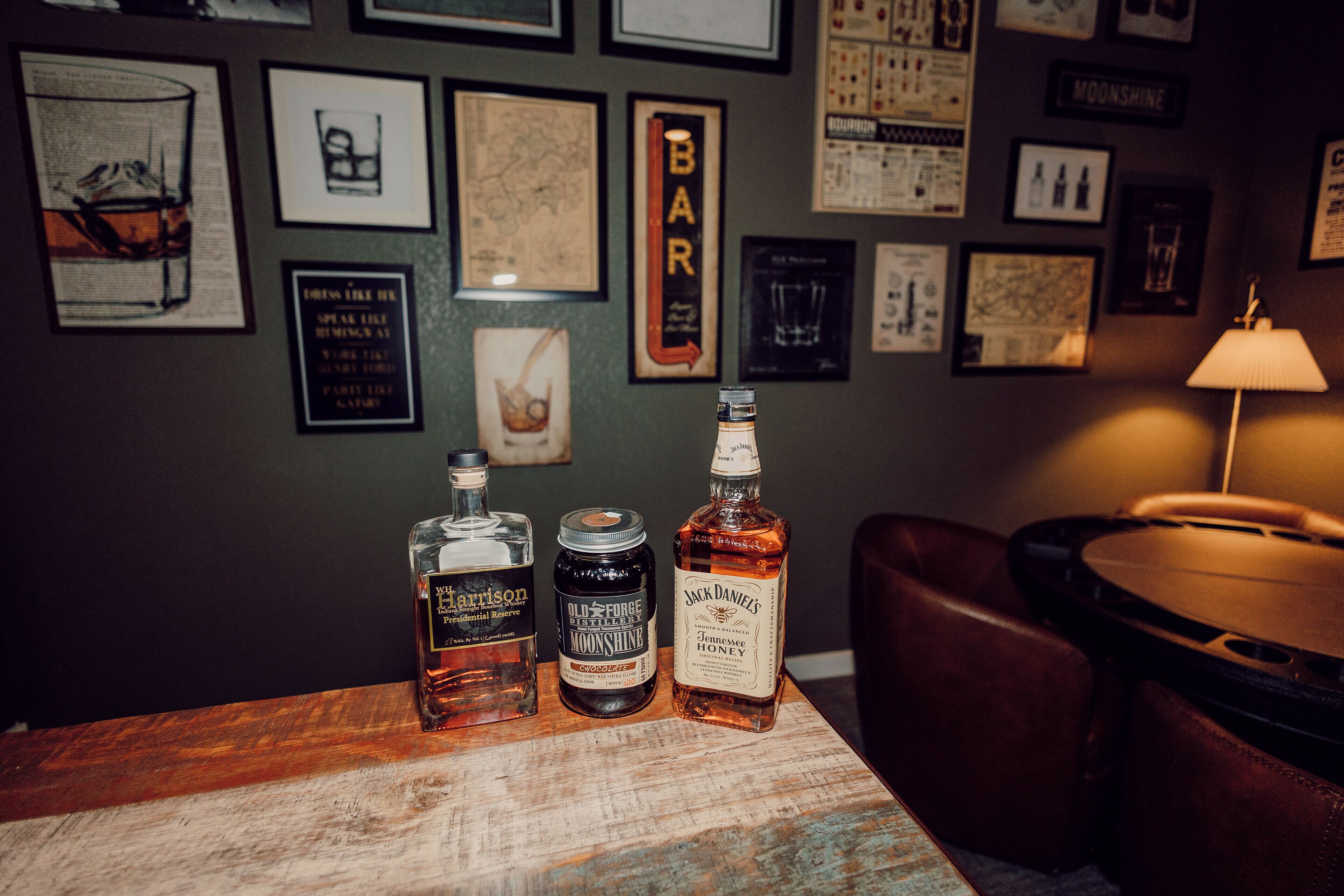 Three bottles of whiskey on a wooden table in a bar setting with framed pictures on the wall.