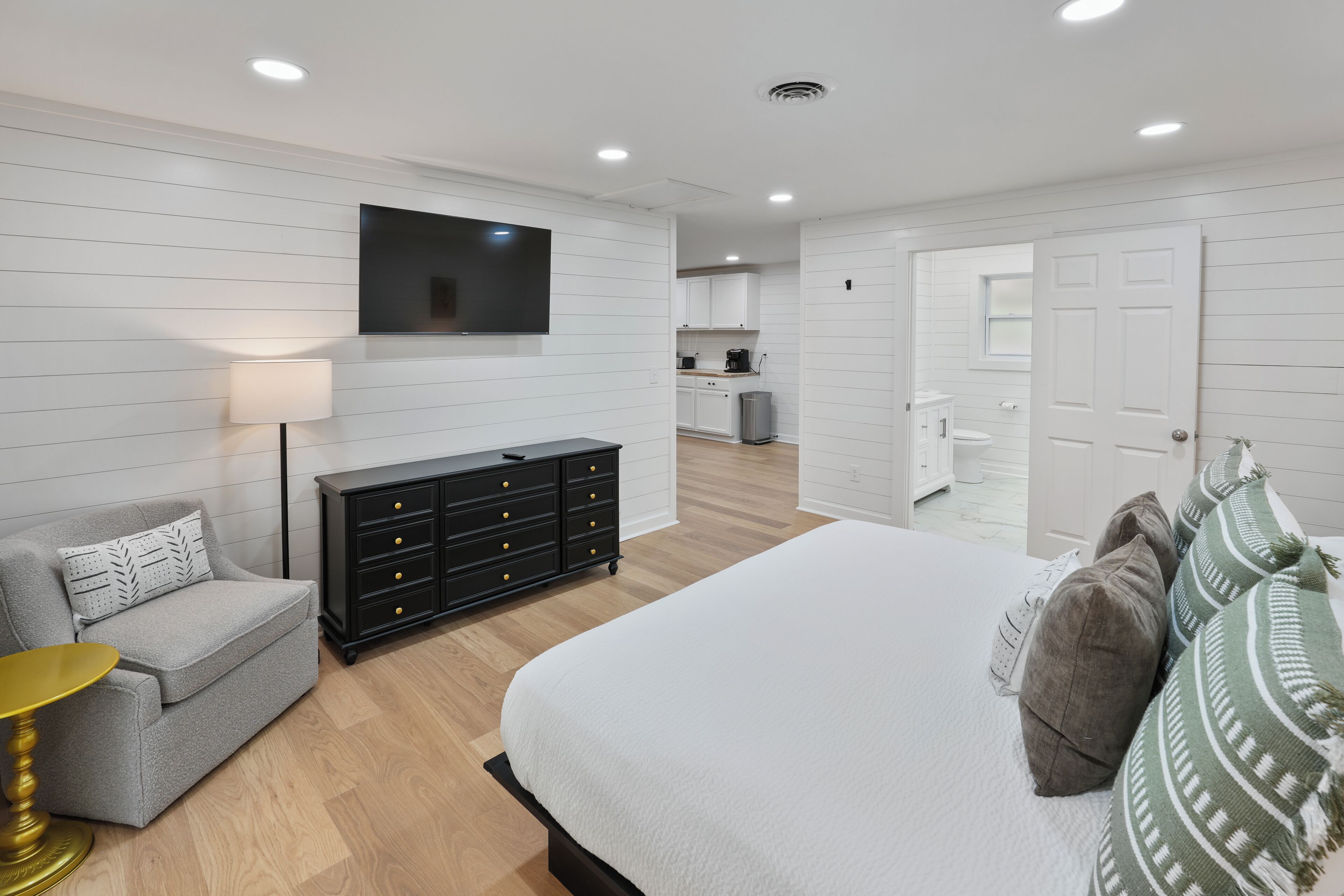 A modern bedroom with a bed, a dresser, a TV, and a kitchenette in the background.