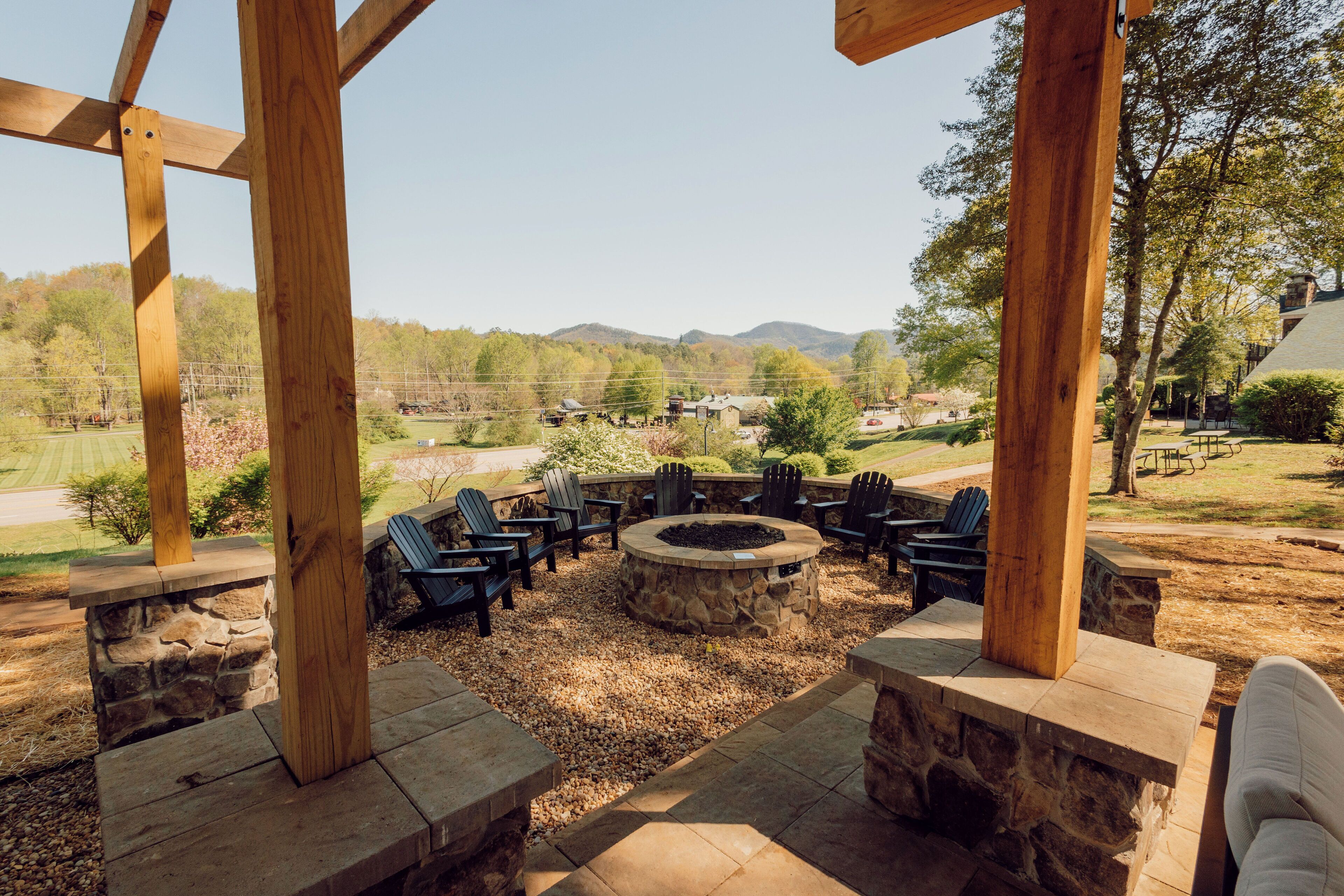 An outdoor seating area with stone fire pit and Adirondack chairs, surrounded by wooden beams and stone walls, overlooking a scenic landscape.