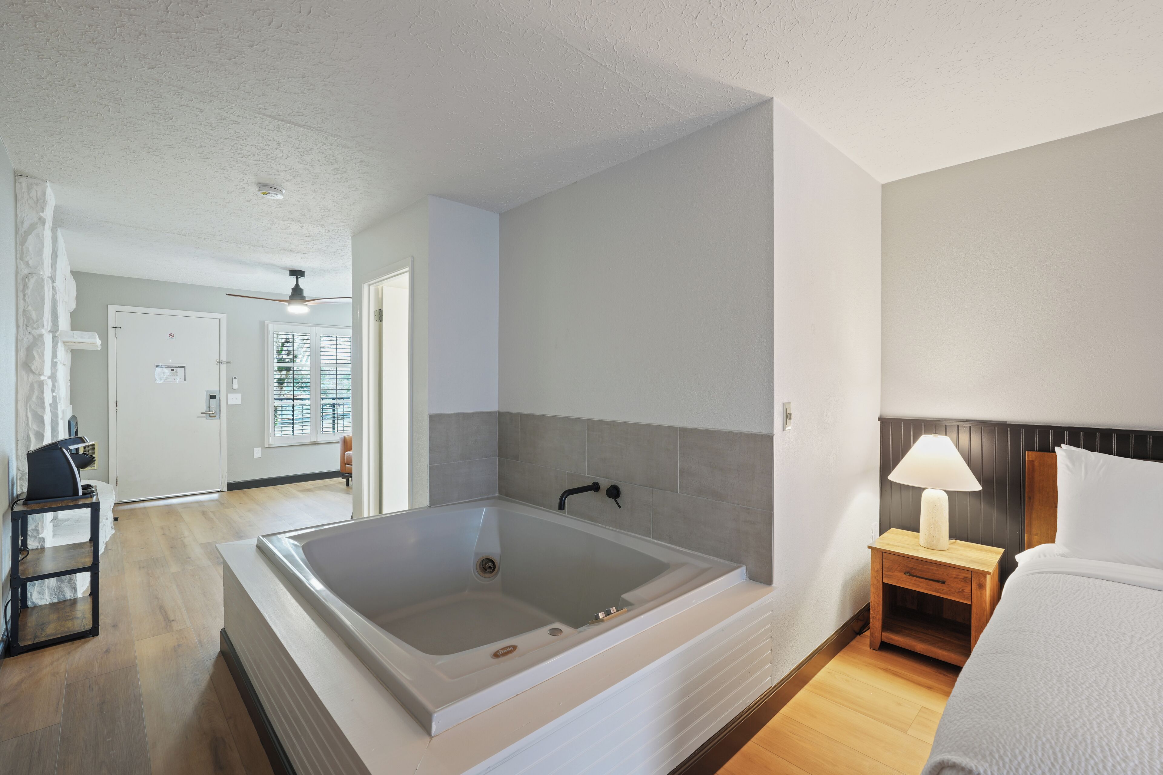 A modern bathroom with a large bathtub, a wooden nightstand with a lamp, and a bed with white bedding.