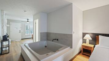 A modern bathroom with a large bathtub, a wooden nightstand with a lamp, and a bed with white bedding.