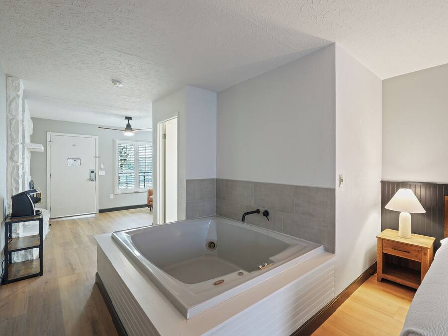 A modern bathroom with a large bathtub, a wooden nightstand with a lamp, and a bed with white bedding.