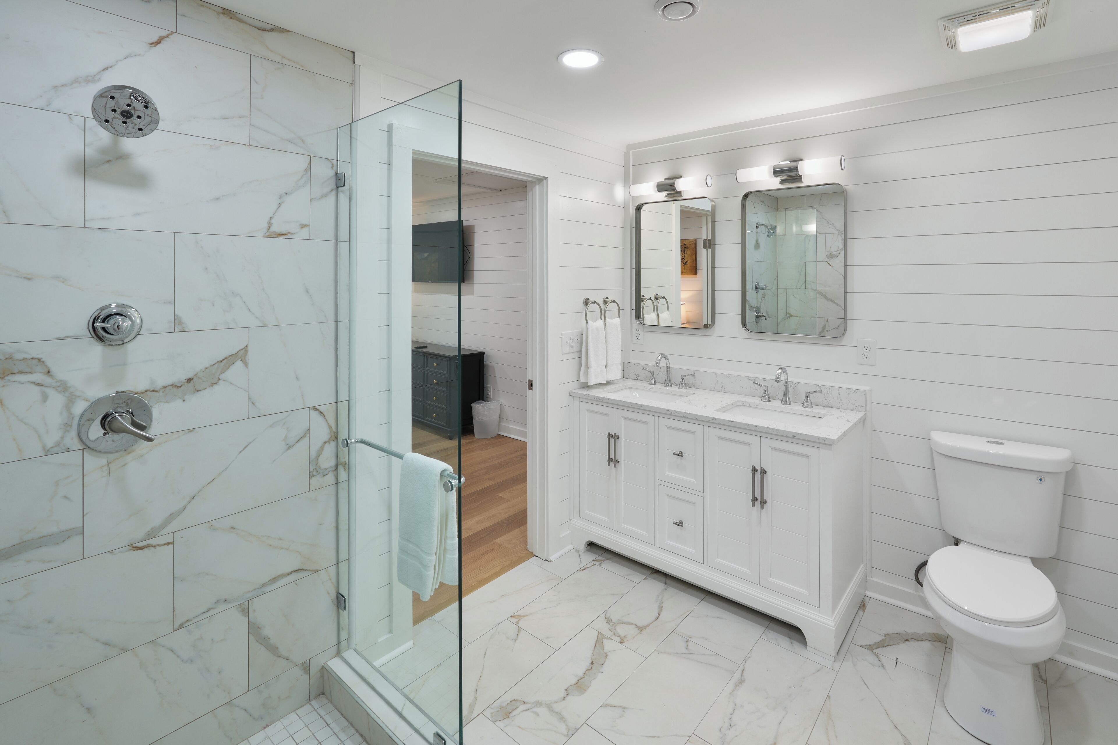A modern bathroom with a glass shower enclosure, white marble tiles, a white sink with a mirror above, and a white toilet.