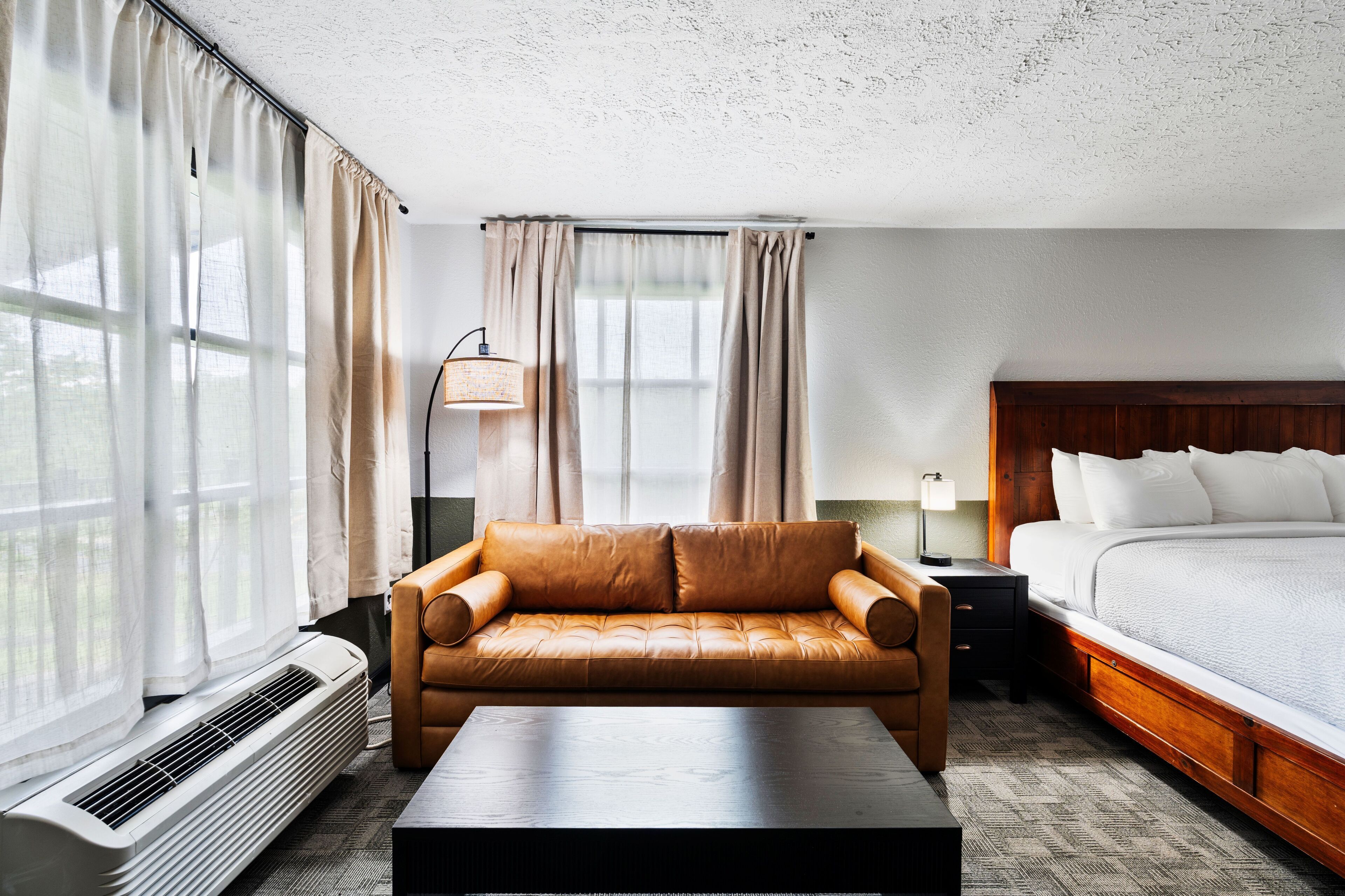 A hotel room with a leather sofa, a bed, a bedside table with a lamp, and a large window with curtains.