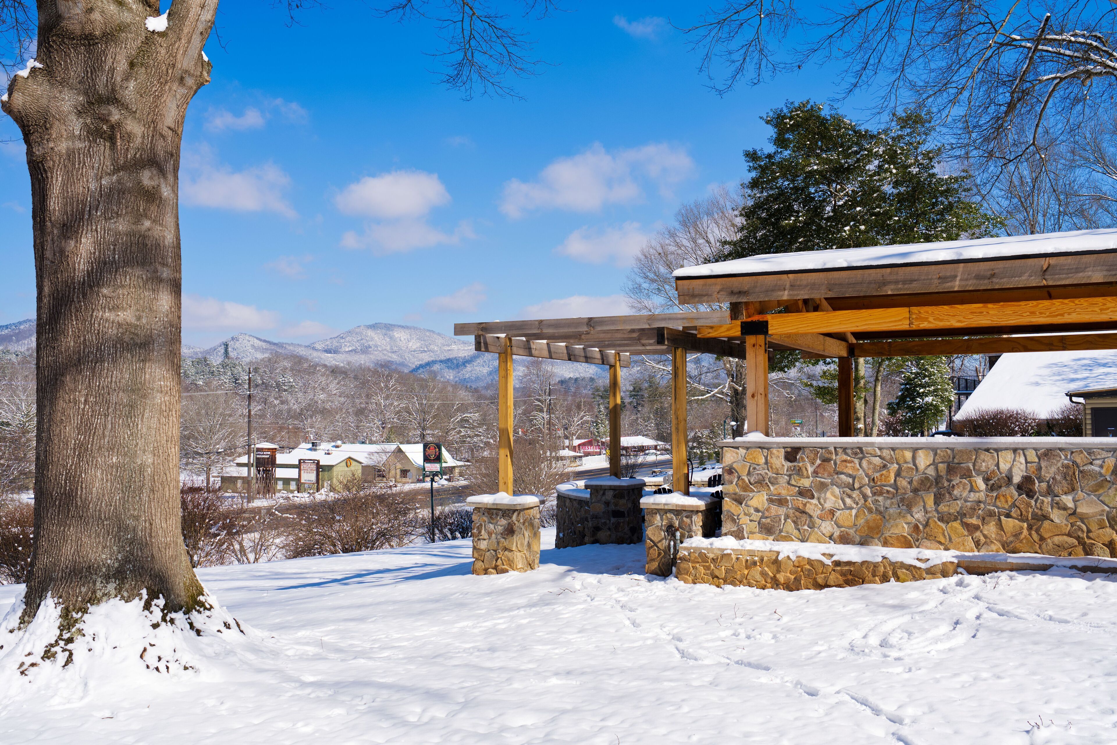 Un espacio exterior cubierto de nieve con un muro de piedra y una pérgola de madera, rodeado de montañas y edificios.