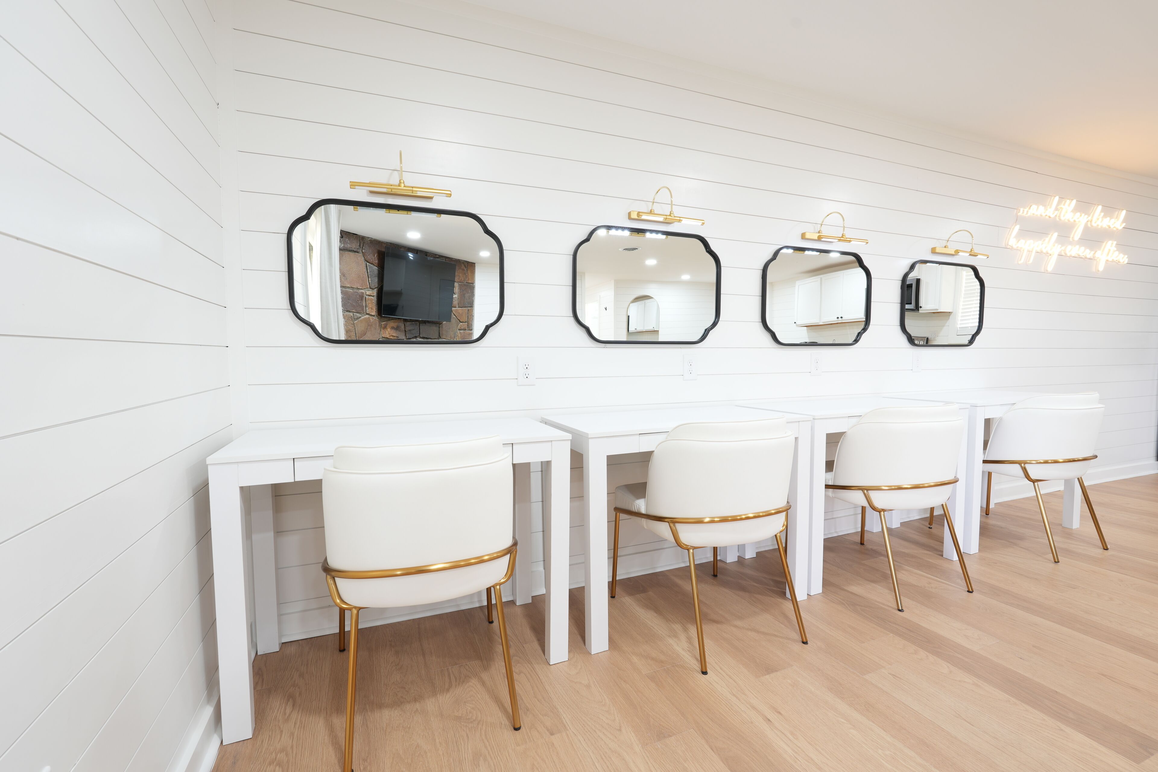 A room with white paneled walls, a wooden floor, and a row of white desks with gold-trimmed chairs.