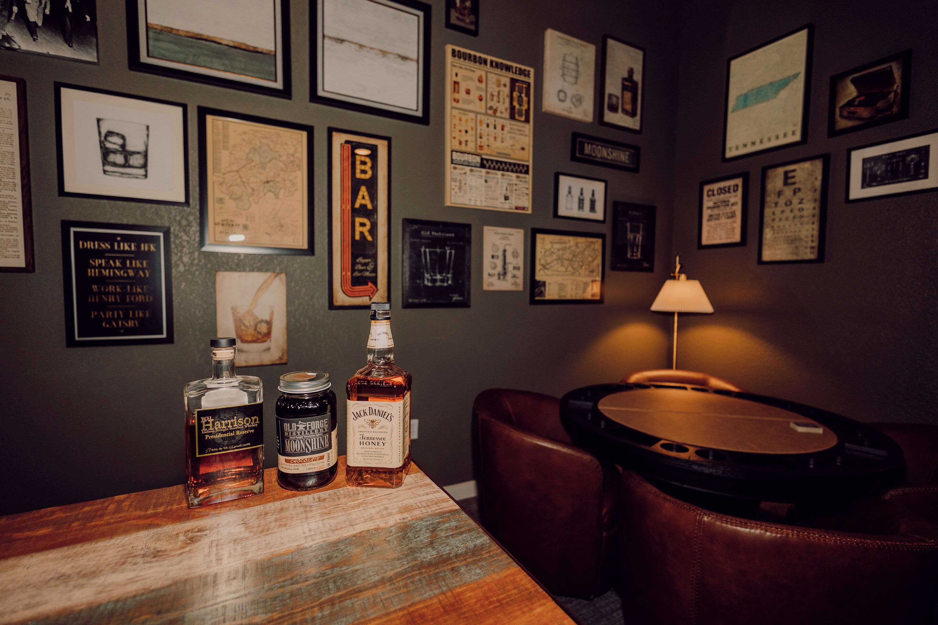 A bar area with a wooden table displaying bottles of whiskey, a sign reading "BAR", and a wall adorned with various framed pictures and posters.