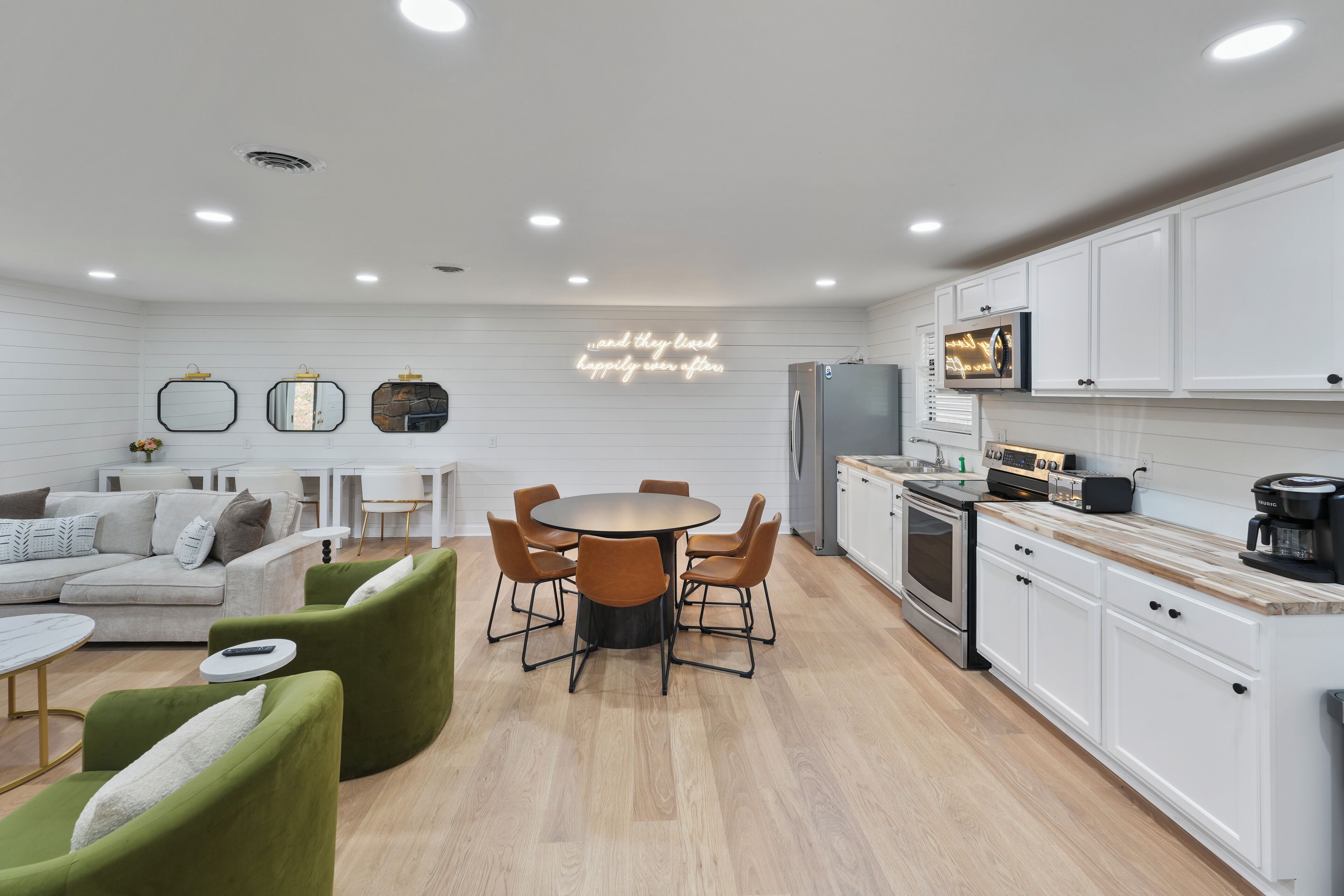 A modern kitchen with white cabinets, a dining area with a round table and chairs, and a living area with a sofa and green armchairs.