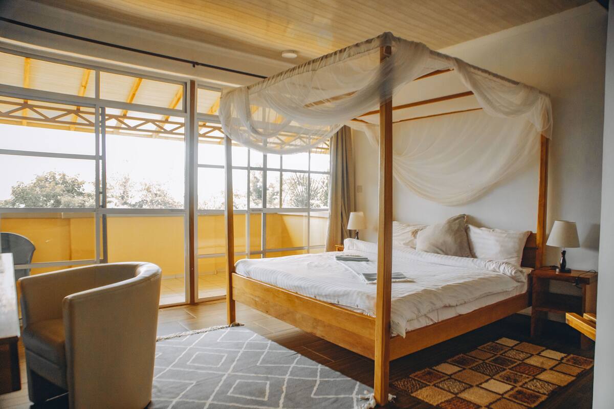 Una cama con dosel, una silla, una mesita de noche con lámpara y vistas al exterior a través de amplios ventanales.