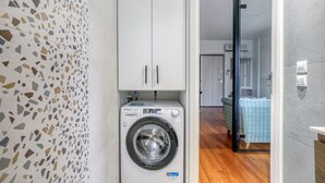 A compact laundry room with a washing machine, a tiled backsplash, and a wooden floor. - Athena's South Apartment (Alimos)