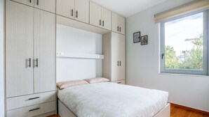 A bedroom with a bed, built-in wardrobe, and a window with a view of greenery. - Athena's South Apartment (Alimos)