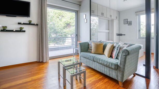 A modern living room with a green sofa, a glass coffee table, wooden flooring, a flat-screen TV, and a sliding glass door leading to a balcony. - Athena's South Apartment (Alimos)