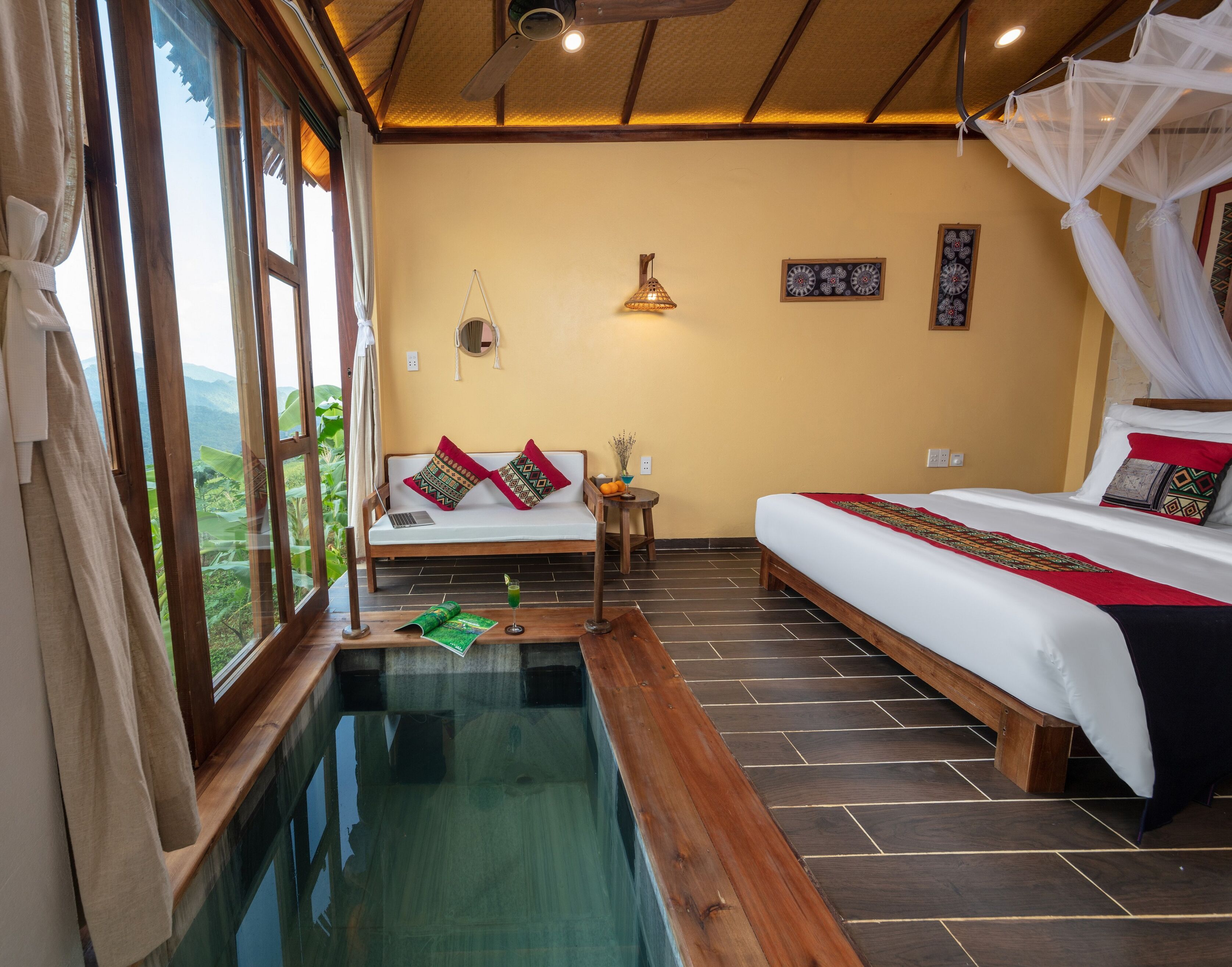Una habitación con cama, sofá, una mesita, una lámpara de noche y vistas a un paisaje exuberante a través de amplios ventanales.