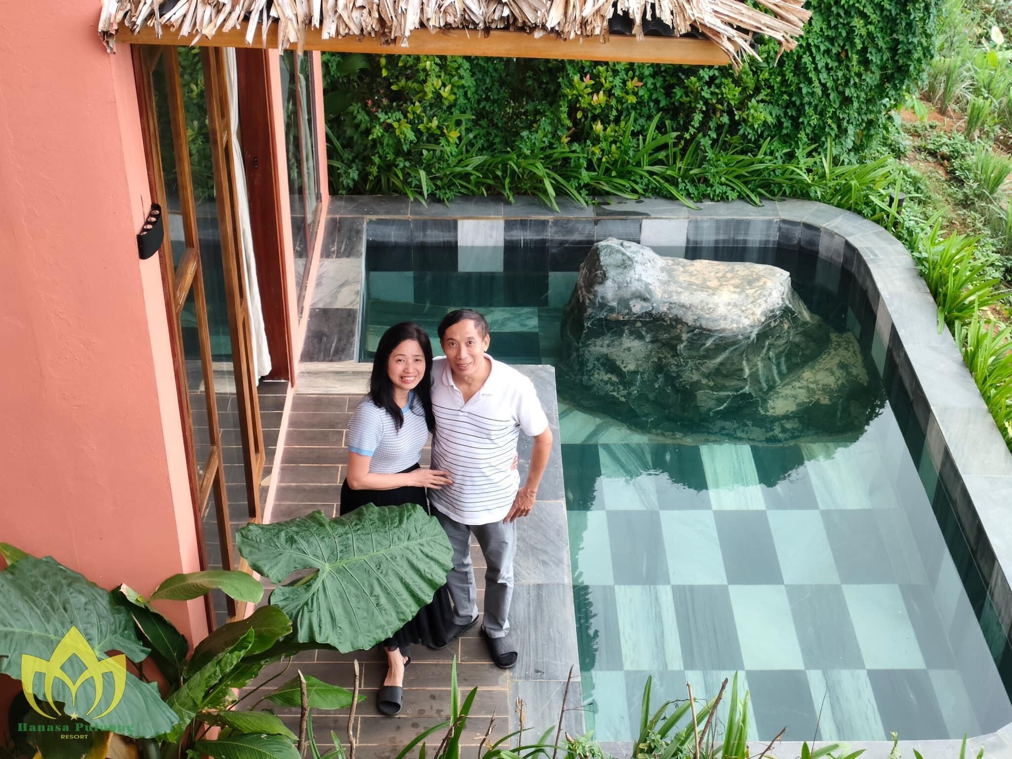 A couple standing by a checkered infinity pool with a large rock, surrounded by lush greenery and a thatched roof structure.