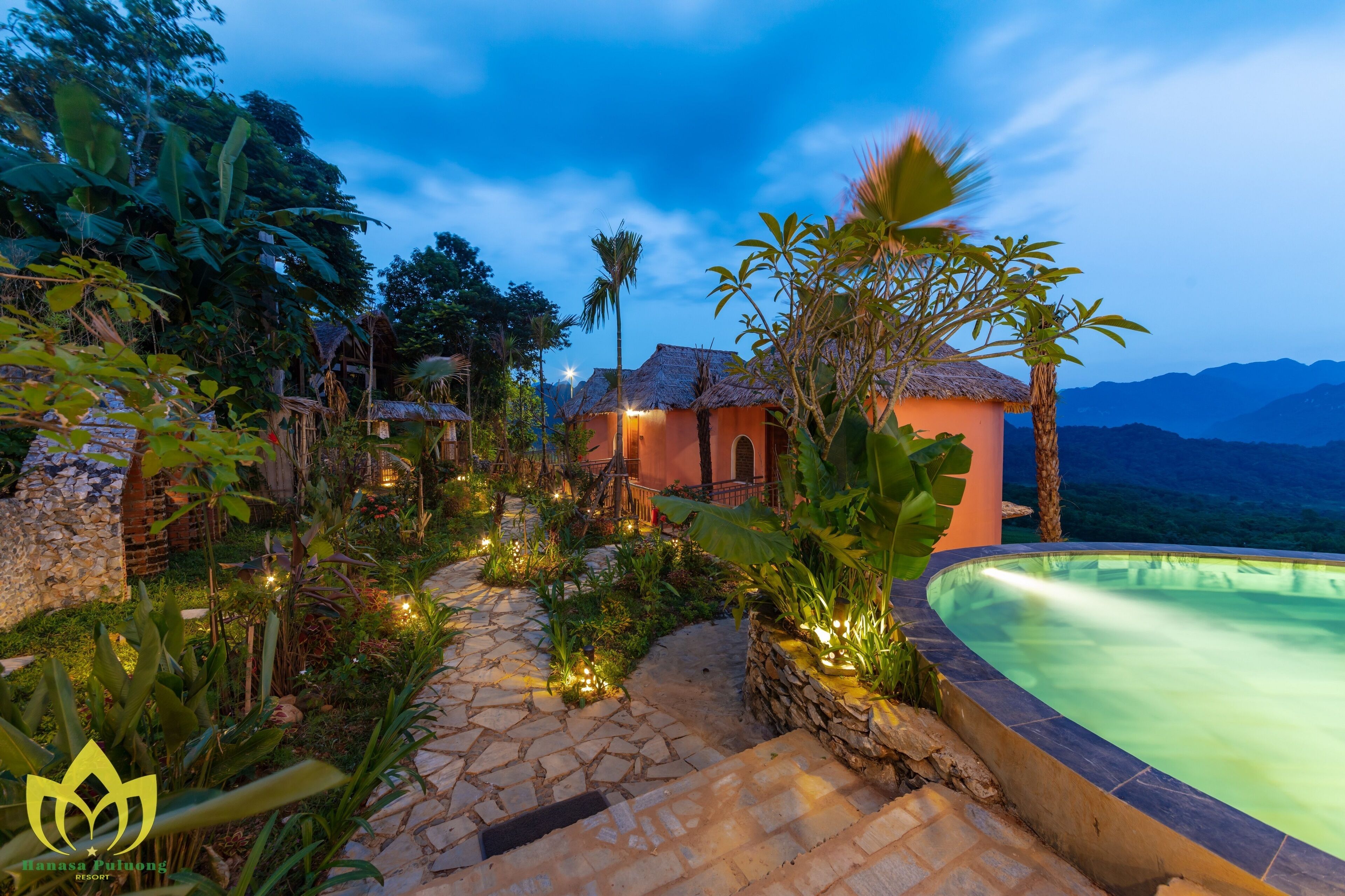 A resort with a stone pathway leading to a pool area, surrounded by tropical plants and a thatched-roof building.
