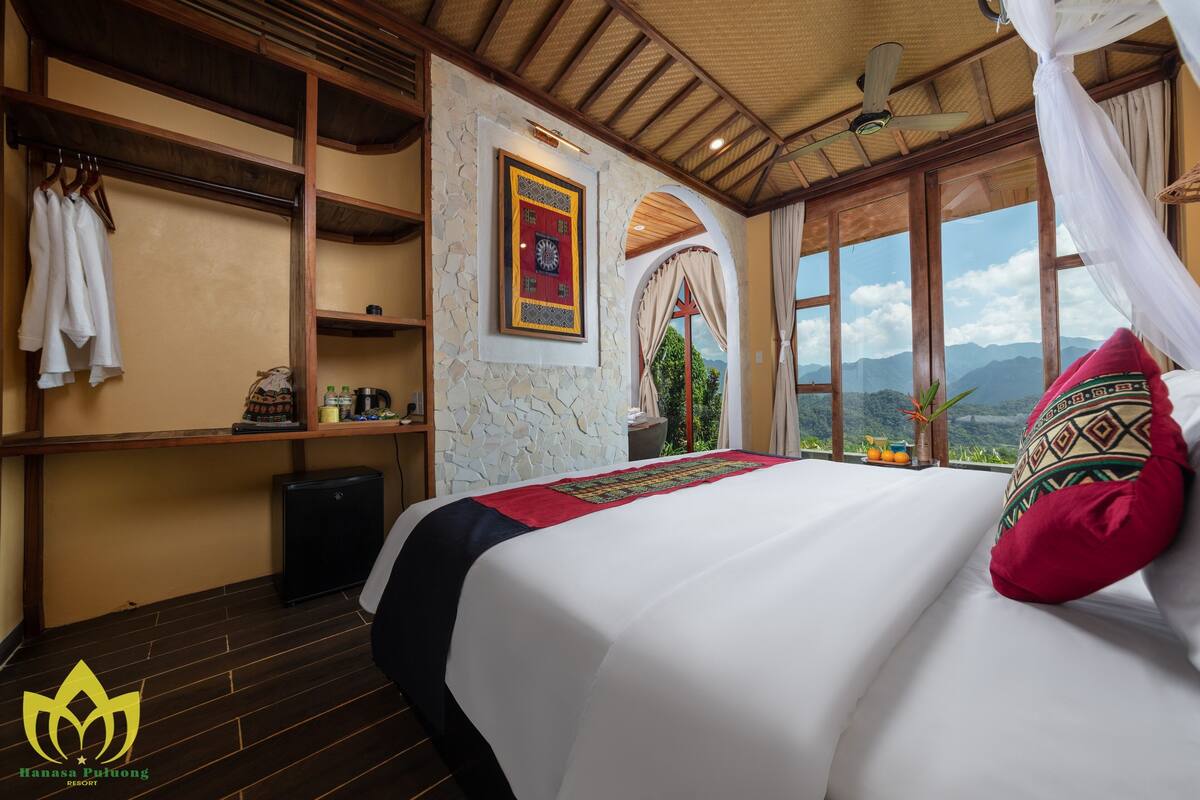 A hotel room with a bed, a clock on the wall, a window with a mountain view, and a ceiling fan.