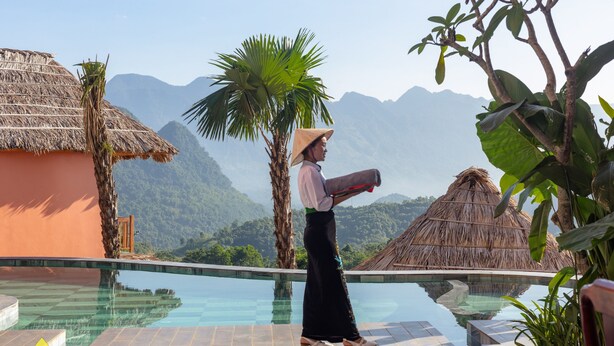 Una persona de pie junto a una piscina con vistas a una montaña, un edificio con techo de paja y plantas tropicales.