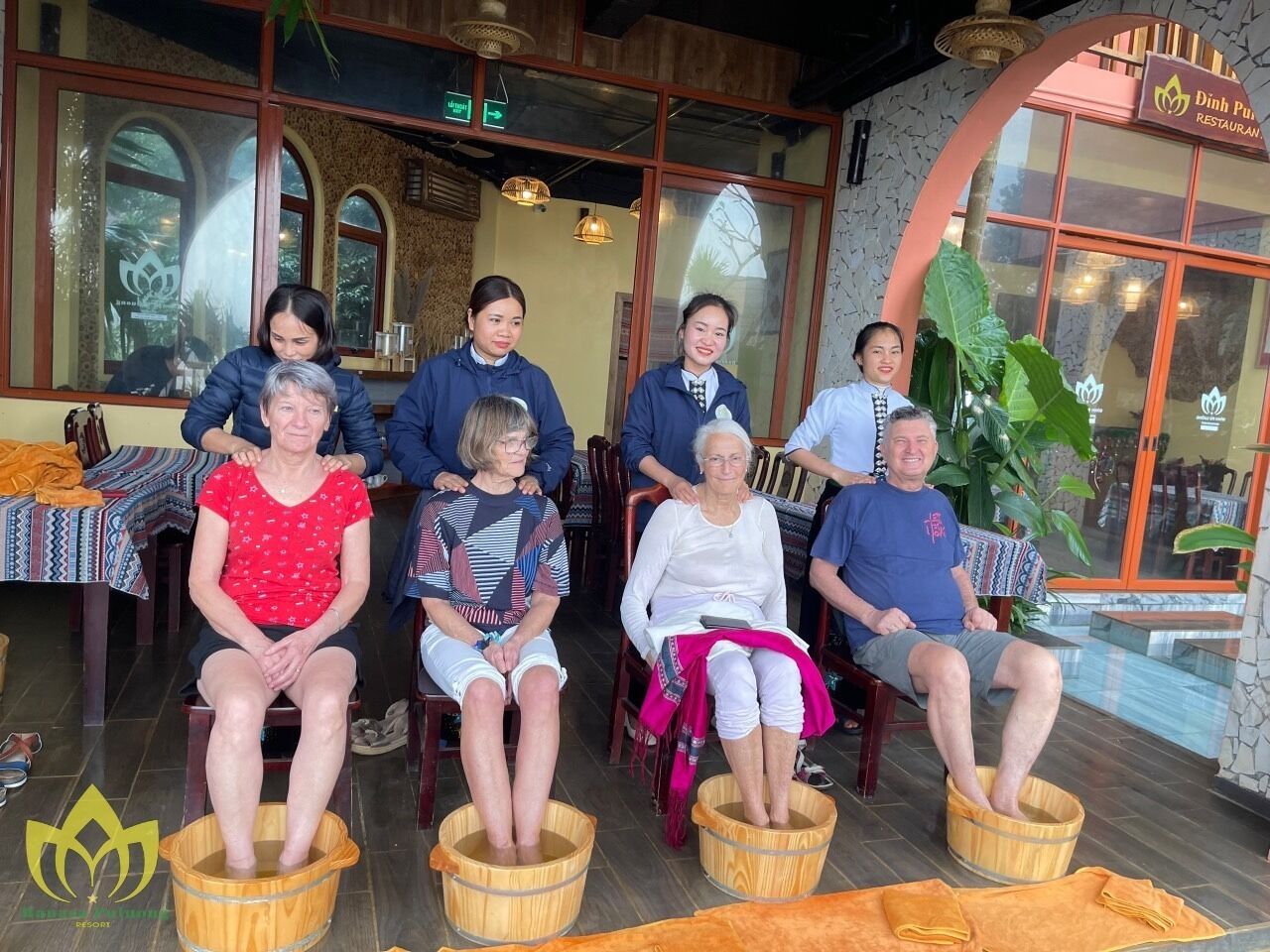 A group of people seated in wooden buckets in a restaurant setting with large windows and indoor plants.
