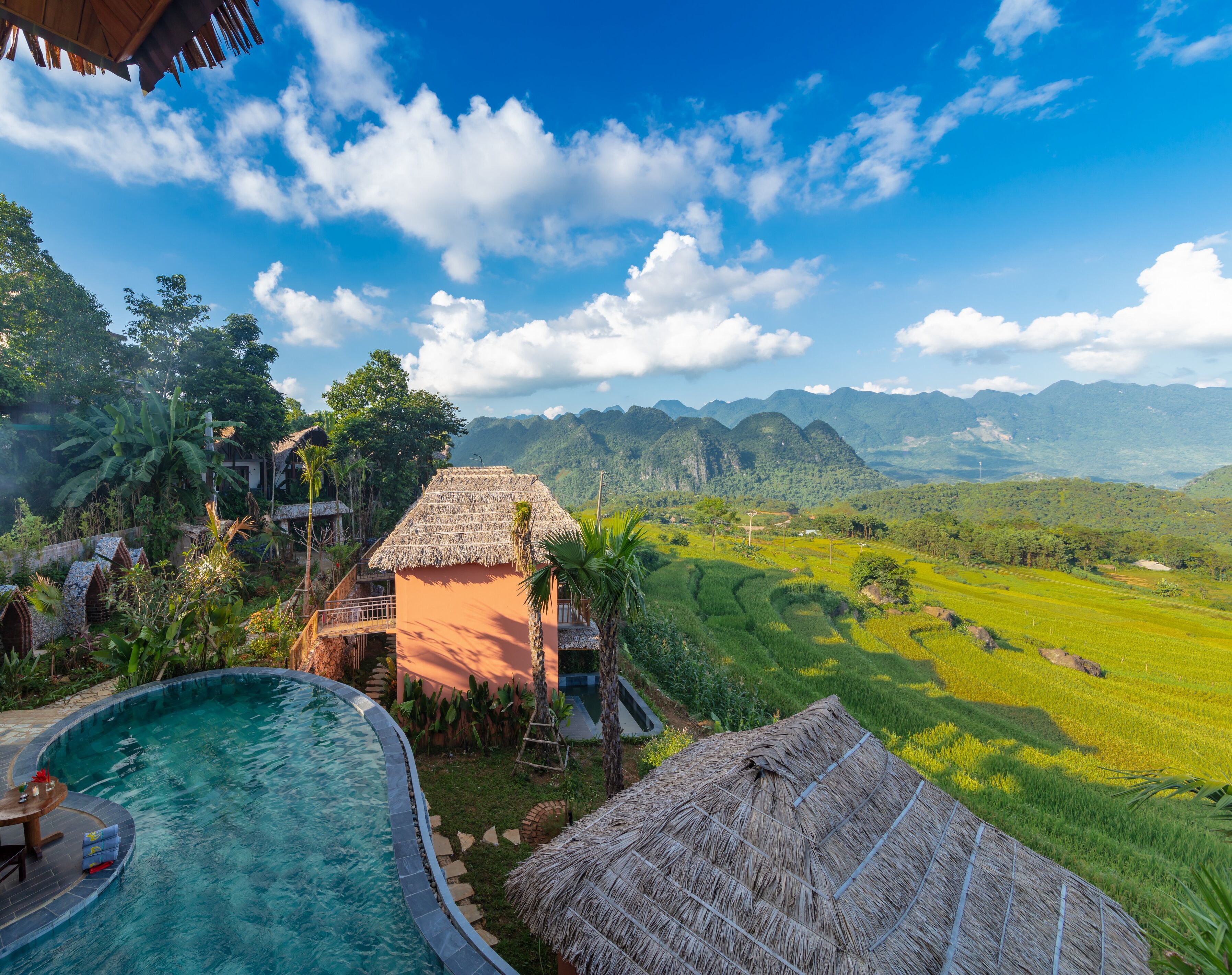 A pool area with a thatched-roof structure, surrounded by lush greenery and a view of distant mountains.