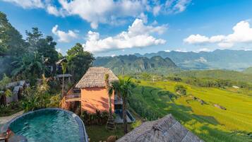 A pool area with a thatched-roof structure, surrounded by lush greenery and a view of distant mountains.