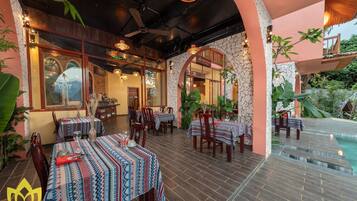 An outdoor restaurant with tables set for dining, surrounded by plants and a pool.